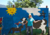 Woman standing outside in front of a large, free-standing mural that depicts a cow, a horse and a child