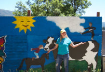 Woman standing outside in front of a large, free-standing mural that depicts a cow, a horse and a child