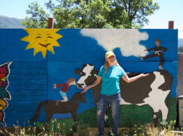Woman standing outside in front of a large, free-standing mural that depicts a cow, a horse and a child