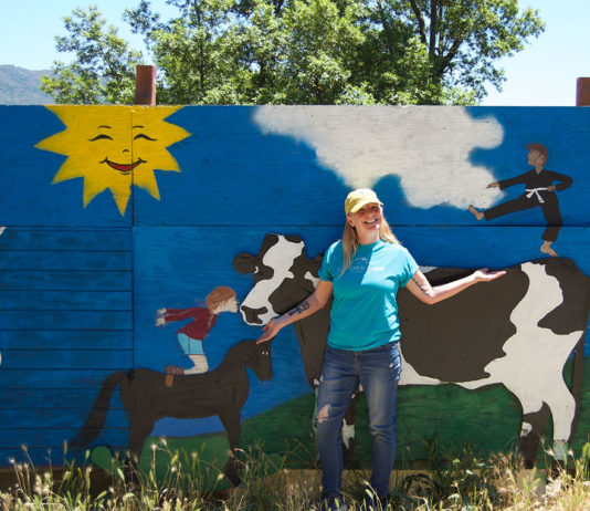 Woman standing outside in front of a large, free-standing mural that depicts a cow, a horse and a child