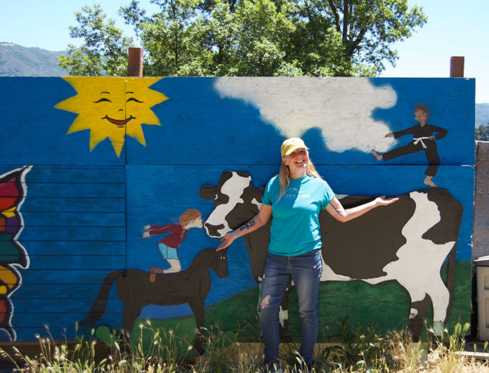 Woman standing outside in front of a large, free-standing mural that depicts a cow, a horse and a child